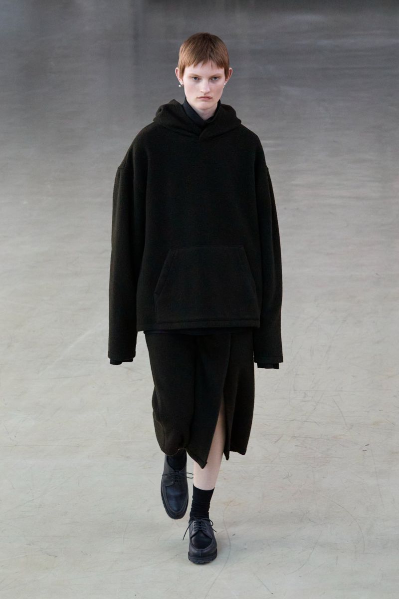 Runway model in black hoodie and skirt walking on a concrete floor, neutral colors, minimalist fashion.