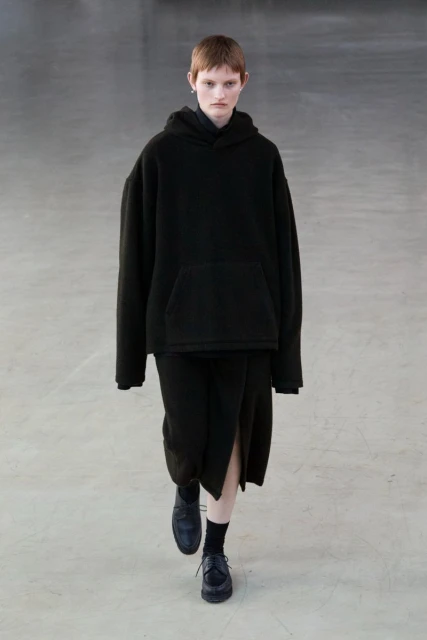 Runway model in black hoodie and skirt walking on a concrete floor, neutral colors, minimalist fashion.