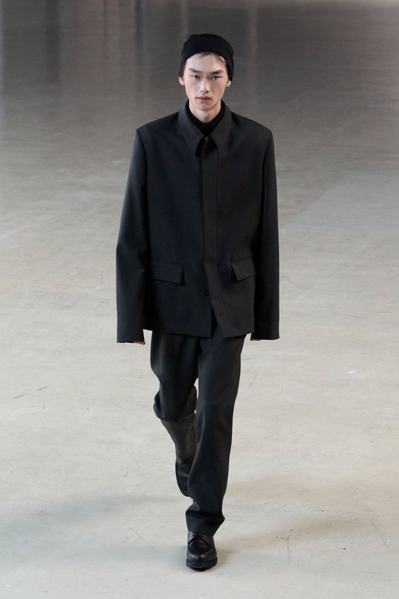 Male fashion model walks on a concrete floor wearing a dark monochrome jacket, pants, shoes, and beanie in a studio setting.