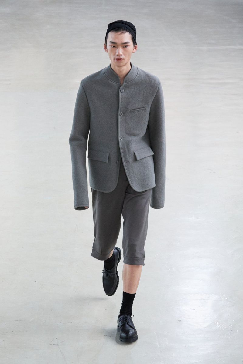 Asian male fashion model walks toward camera on a light-colored seamless backdrop, wearing a gray jacket, gray shorts, black socks, and black shoes.
