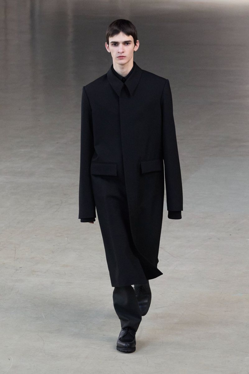 Male model in a black coat and pants walks a neutral runway, showcasing fashion design with simple background and natural light.