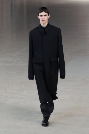Male model in a black coat and pants walks a neutral runway, showcasing fashion design with simple background and natural light.