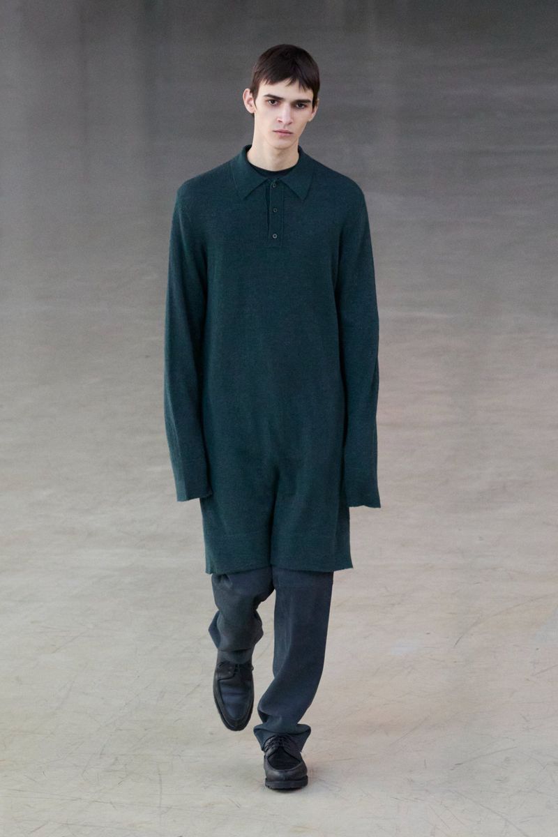 Model wearing a dark green long-sleeved polo tunic and dark trousers walking forward on a neutral grey concrete floor