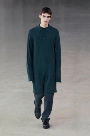 Model wearing a dark green long-sleeved polo tunic and dark trousers walking forward on a neutral grey concrete floor