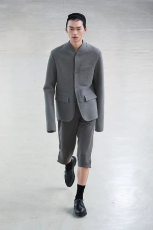 Model wearing grey jacket and dark shorts walking forward on a neutral concrete floor