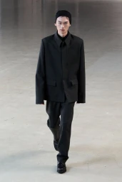 A model walking on a concrete floor wearing a dark jacket, dark trousers, and a black beanie for the Rier F/W 25 lookbook.