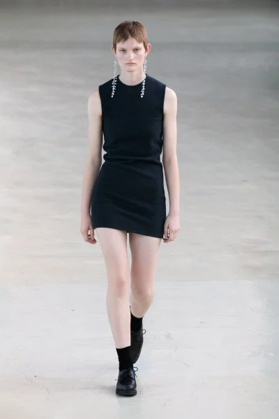 Model wearing a black sleeveless dress and drop earrings walking forward on a neutral floor