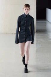 Model walking forward in a dark button-down minidress and black shoes on a glossy concrete floor.