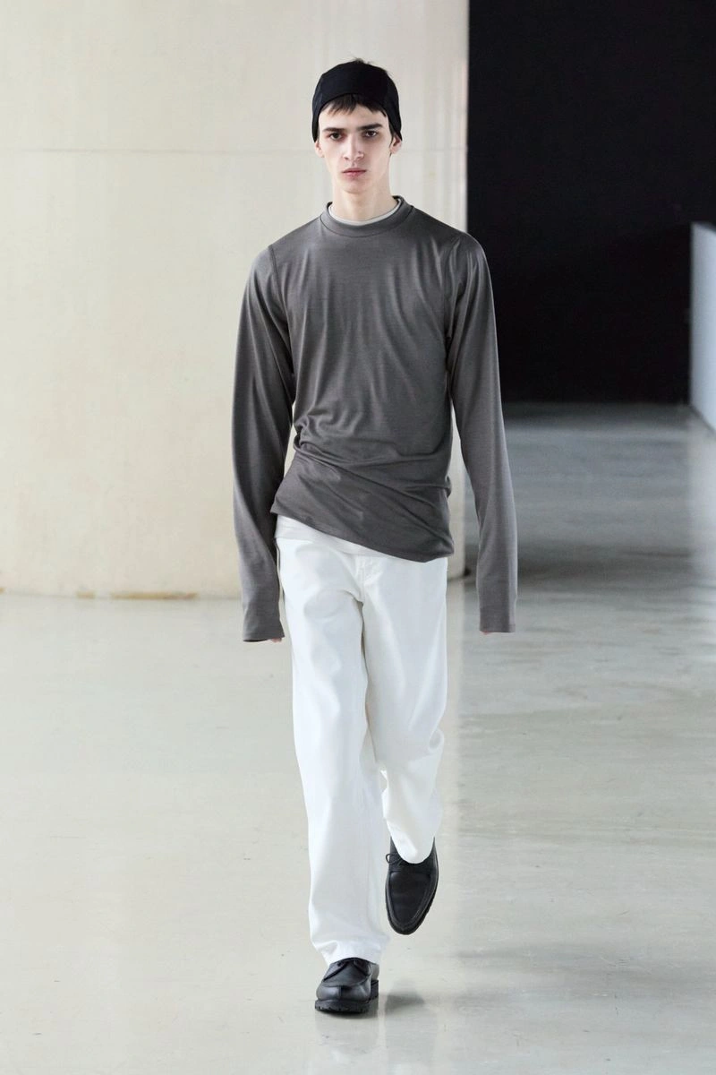 Model wearing a grey long-sleeve top, white trousers, and a black beanie walking in a modern architectural interior.