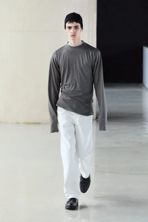 Model wearing a grey long-sleeve top, white trousers, and a black beanie walking in a modern architectural interior.