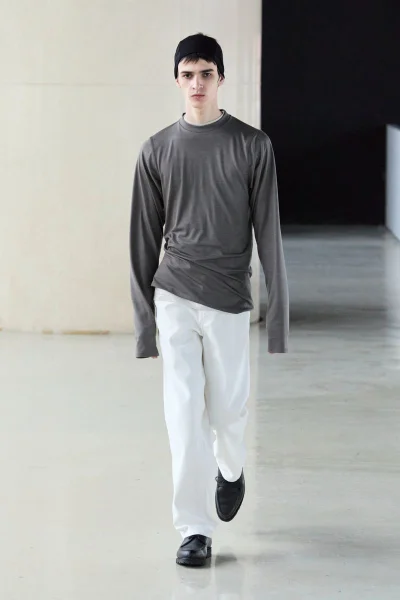 Model wearing a grey long-sleeve top, white trousers, and a black beanie walking in a modern architectural interior.