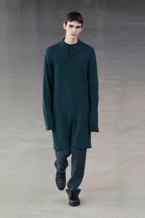 Model wearing a dark green long-sleeved polo tunic and dark trousers walking forward on a neutral grey concrete floor