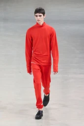 Model walking a runway wearing a red turtleneck top and matching red trousers against a grey floor