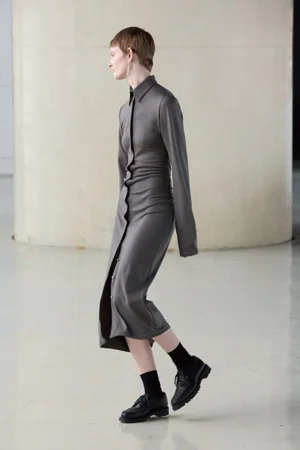 Model in a long grey button-down shirt dress with black socks and shoes walking against a light architectural background.