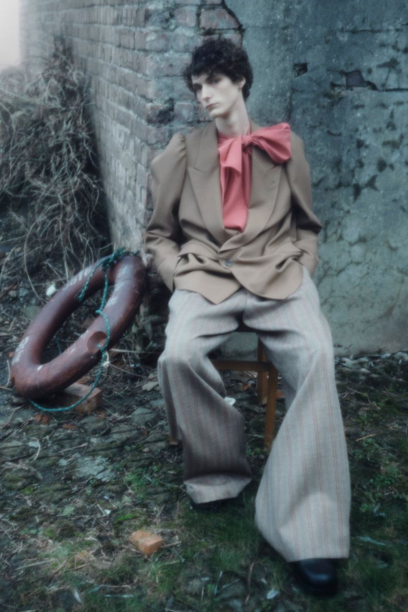 Adult male fashion model in beige suit with pink bow sits on wooden chair in outdoor setting with brick wall and life preserver.
