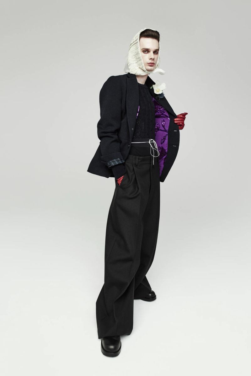 Full-length studio shot of a person in a black jacket, wide-leg pants, headscarf, and gloves, posing in front of a gray background.