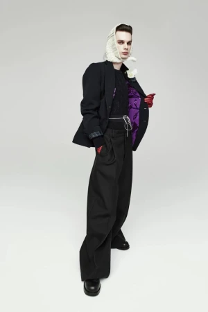 Full-length studio shot of a person in a black jacket, wide-leg pants, headscarf, and gloves, posing in front of a gray background.