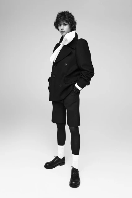 Black and white portrait of a young adult wearing a double-breasted jacket, shorts, stockings, socks, and shoes against a plain background.