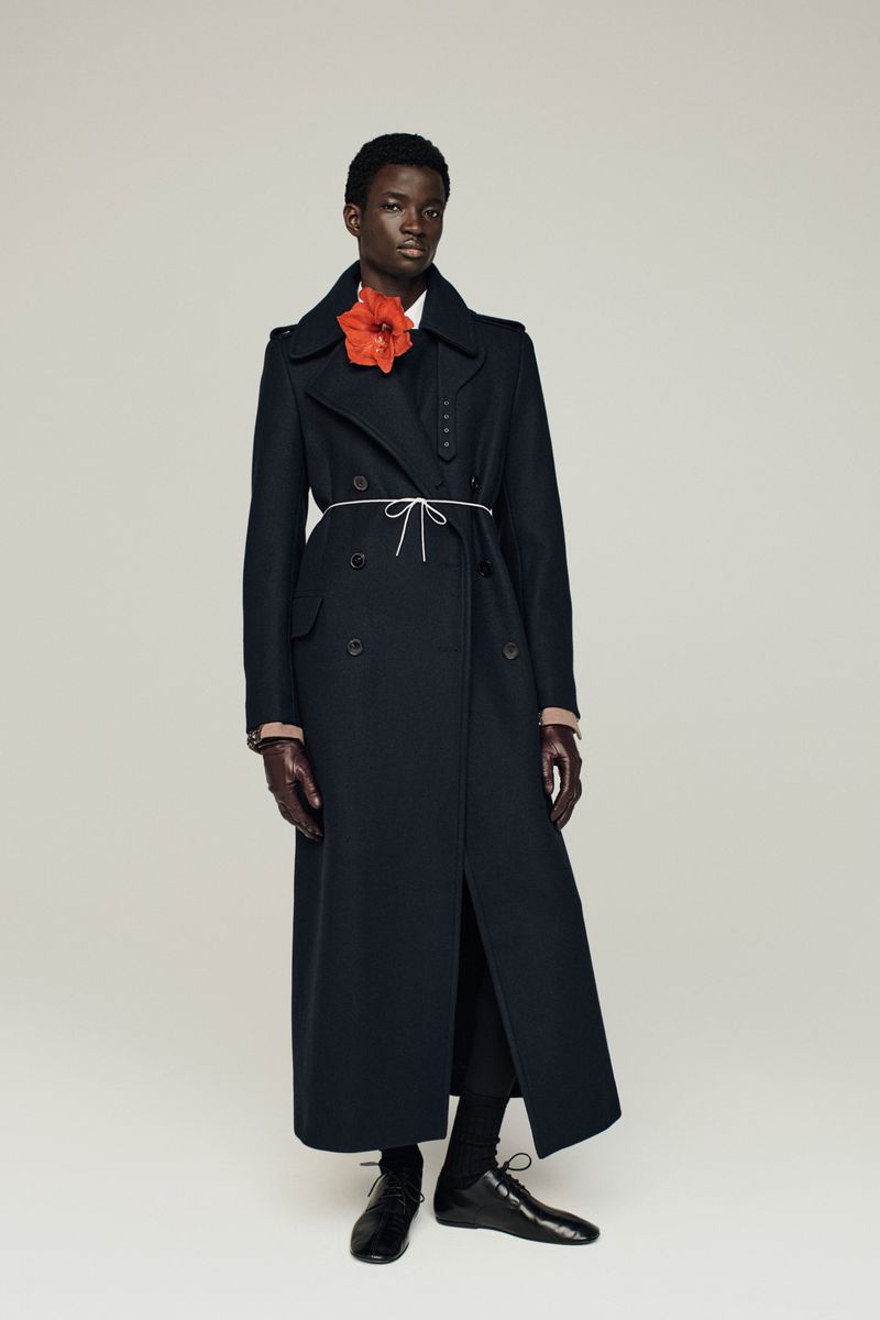Dark-skinned model wears long black coat and flower on a light background in a studio shot, showcasing fashion and style.