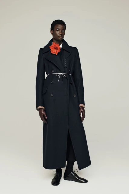 Dark-skinned model wears long black coat and flower on a light background in a studio shot, showcasing fashion and style.