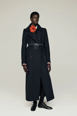Dark-skinned model wears long black coat and flower on a light background in a studio shot, showcasing fashion and style.
