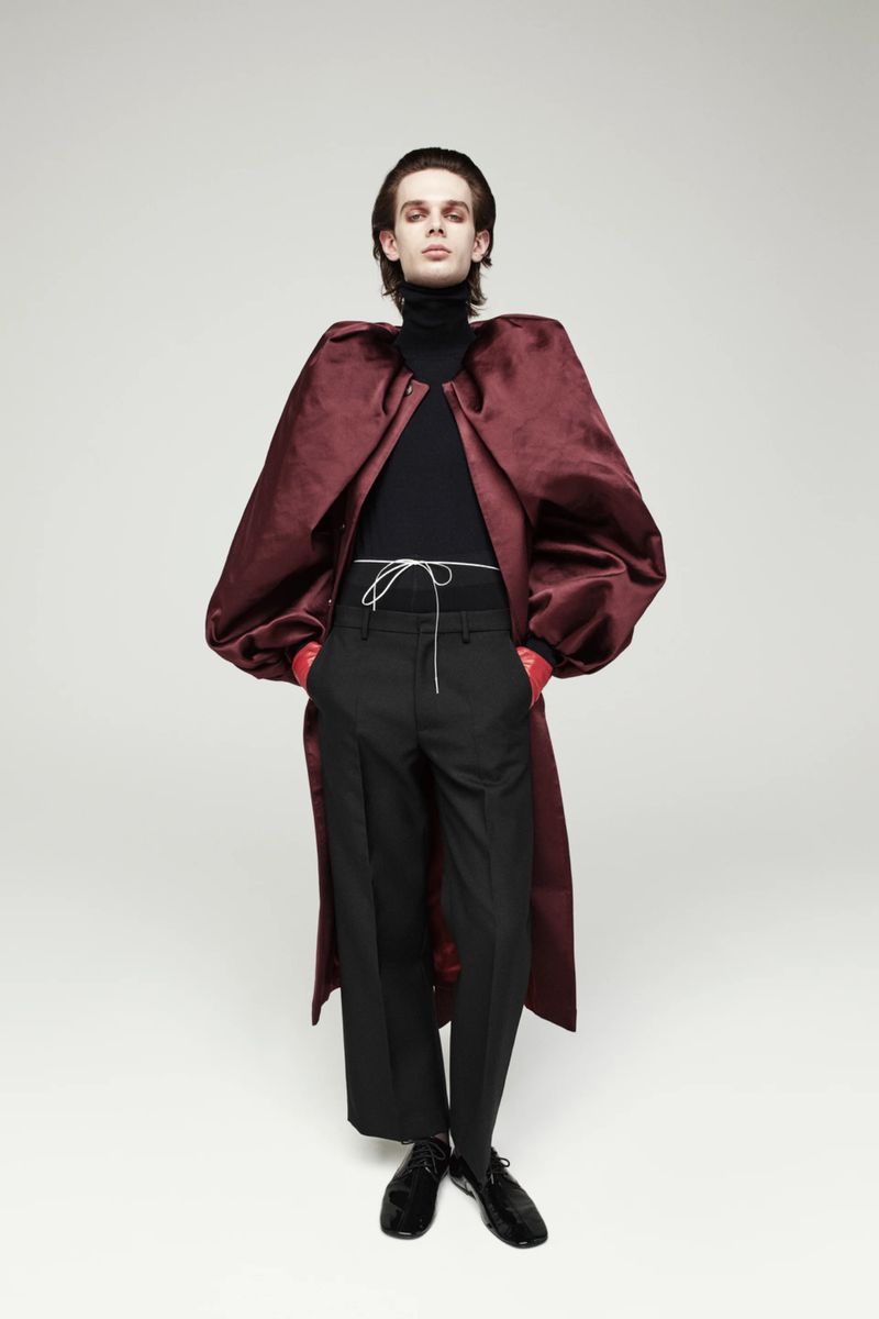 A slender person poses in a black turtleneck, black pants, and a burgundy velvet coat in a full shot against a neutral background.