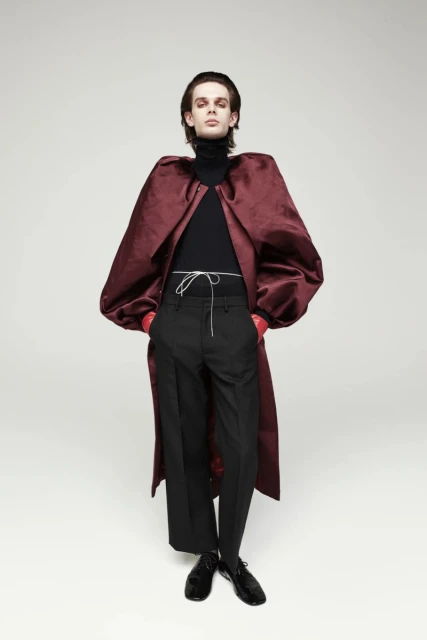 A slender person poses in a black turtleneck, black pants, and a burgundy velvet coat in a full shot against a neutral background.