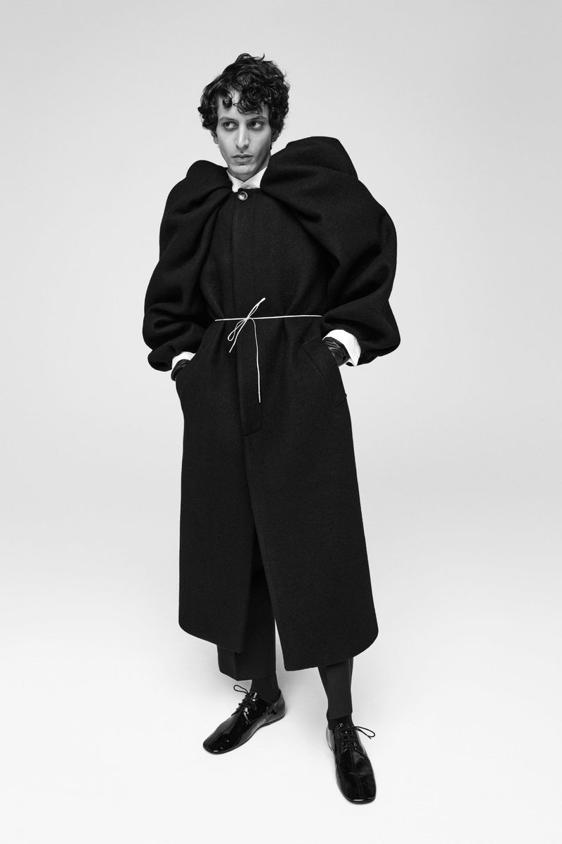 A person with dark curly hair posing in a black coat cinched at the waist with a string, paired with wide-leg trousers and patent leather shoes.