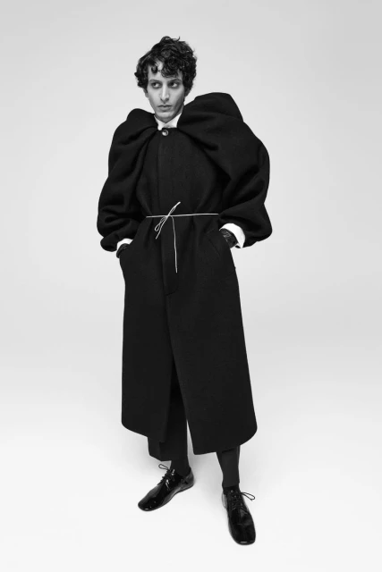 A person with dark curly hair posing in a black coat cinched at the waist with a string, paired with wide-leg trousers and patent leather shoes.