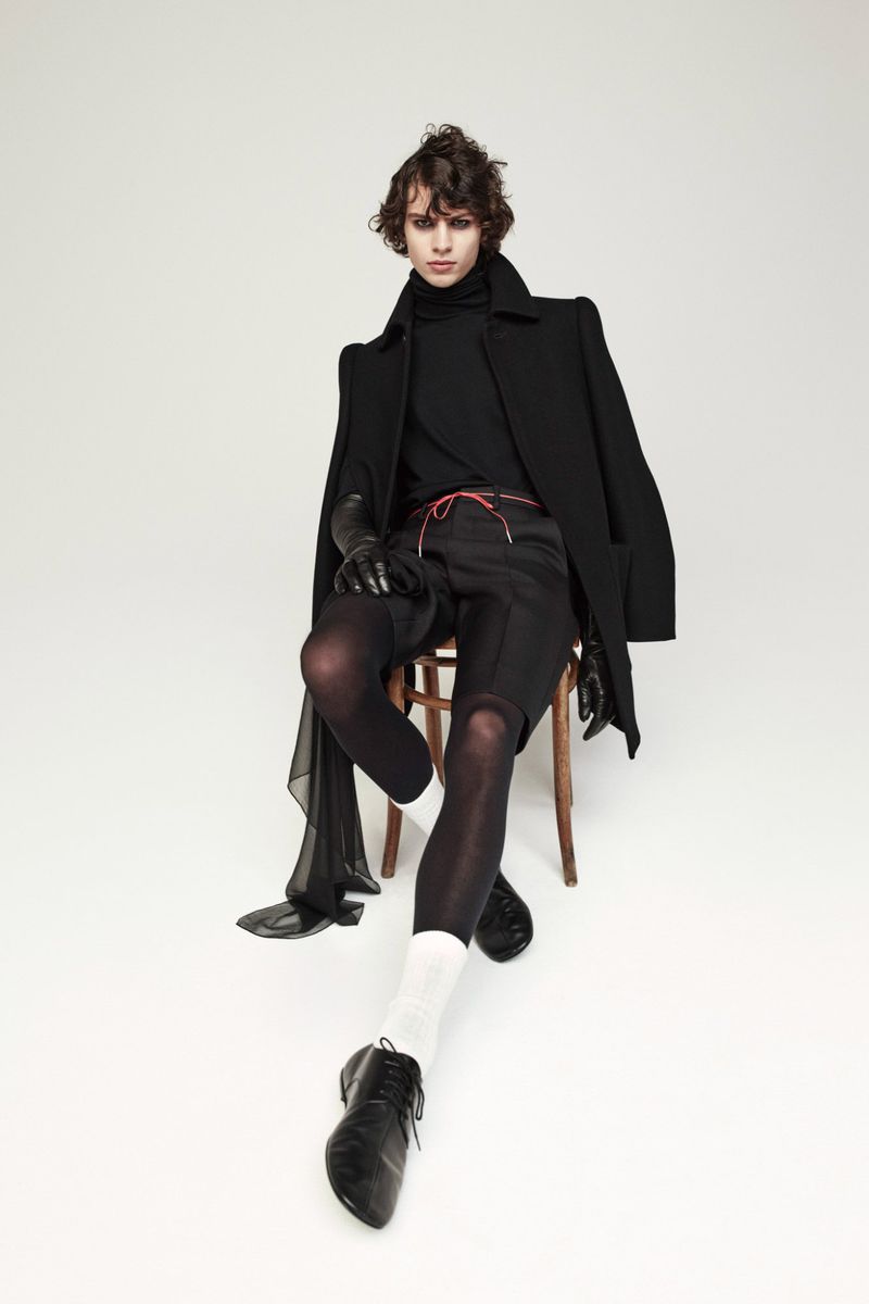 A young adult with curly hair sits on a wooden chair modeling a black coat, black turtleneck, dark shorts, and black leather dress shoes.