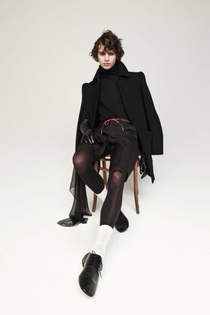 A young adult with curly hair sits on a wooden chair modeling a black coat, black turtleneck, dark shorts, and black leather dress shoes.