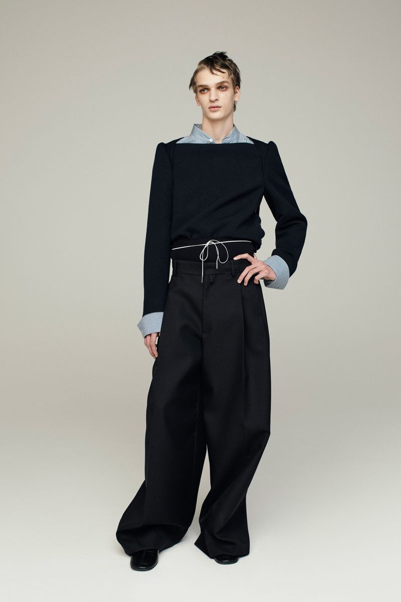 Fashion portrait of a young adult with striking eye makeup, wearing a black top, light-blue collared shirt, and wide-leg black trousers indoors.