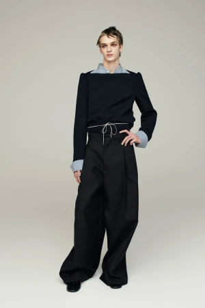 Fashion portrait of a young adult with striking eye makeup, wearing a black top, light-blue collared shirt, and wide-leg black trousers indoors.