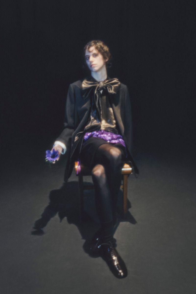 A person sits on a chair in a dark studio setting, wearing a black coat and shiny shoes while holding a flower in hand.