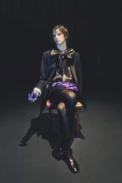 A person sits on a chair in a dark studio setting, wearing a black coat and shiny shoes while holding a flower in hand.