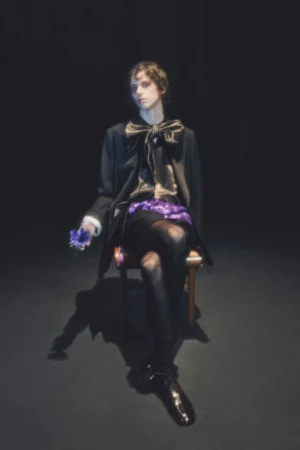 A person sits on a chair in a dark studio setting, wearing a black coat and shiny shoes while holding a flower in hand.