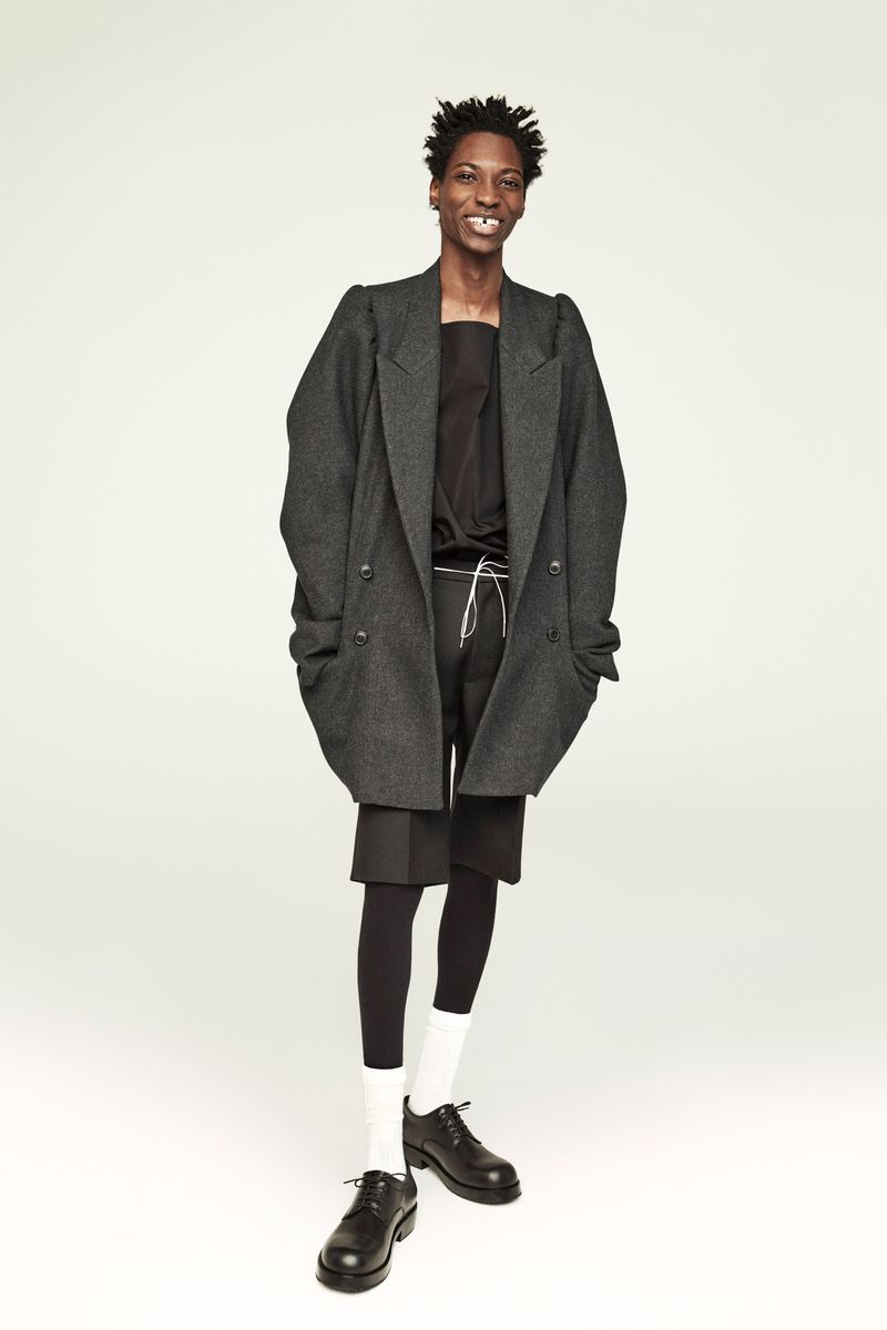 Full-length portrait of a smiling Black male model in a grey wool coat, black top and shorts, leggings, white socks, and black leather shoes.