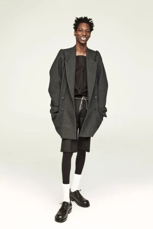 Full-length portrait of a smiling Black male model in a grey wool coat, black top and shorts, leggings, white socks, and black leather shoes.