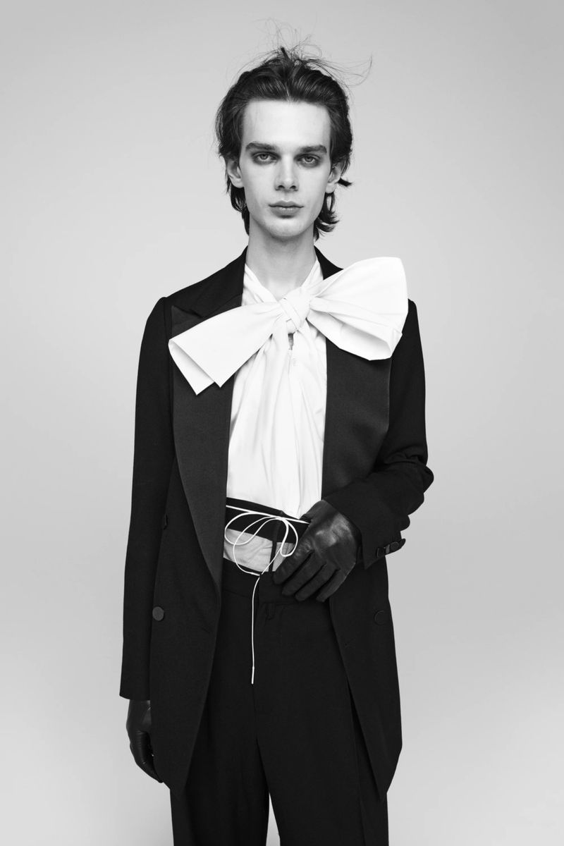 Monochrome studio portrait of a slender adult male wearing a black tuxedo jacket and pants with a large white bow and leather gloves.