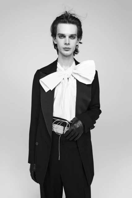 Monochrome studio portrait of a slender adult male wearing a black tuxedo jacket and pants with a large white bow and leather gloves.