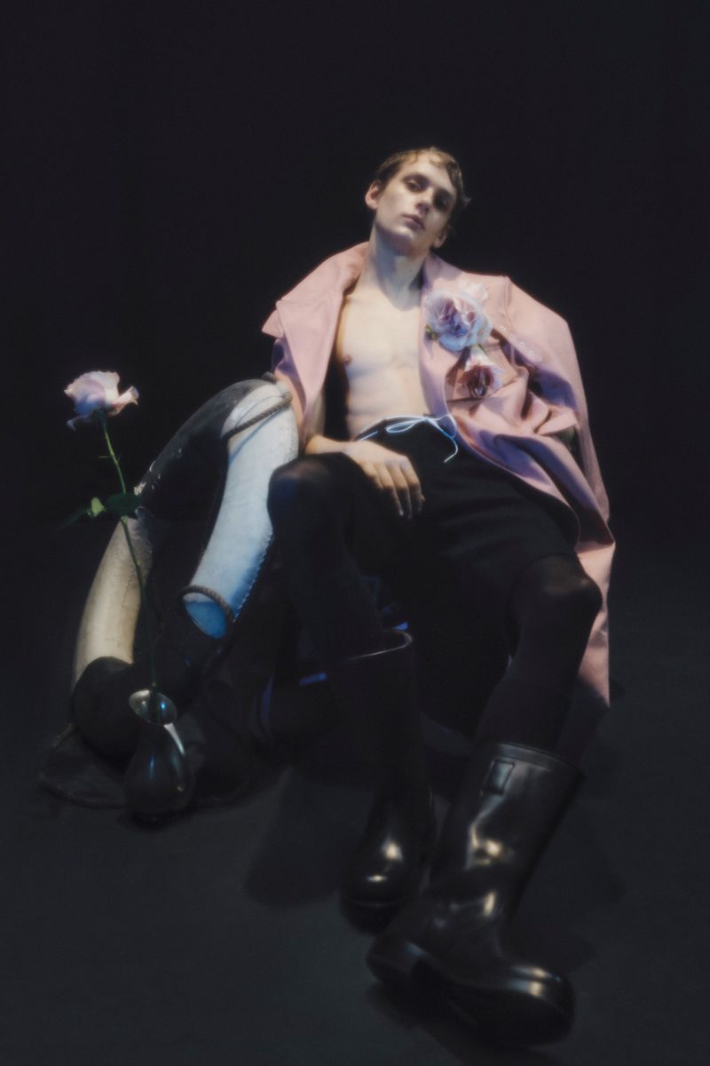 Young man poses with bare torso, wearing a pink coat, black pants and boots, seated by floral arrangement on a black background.