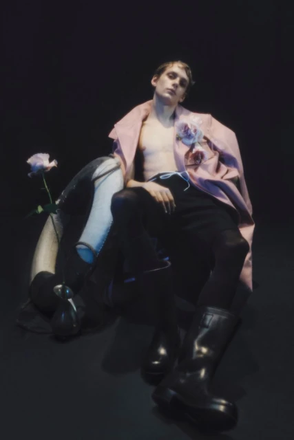 Young man poses with bare torso, wearing a pink coat, black pants and boots, seated by floral arrangement on a black background.