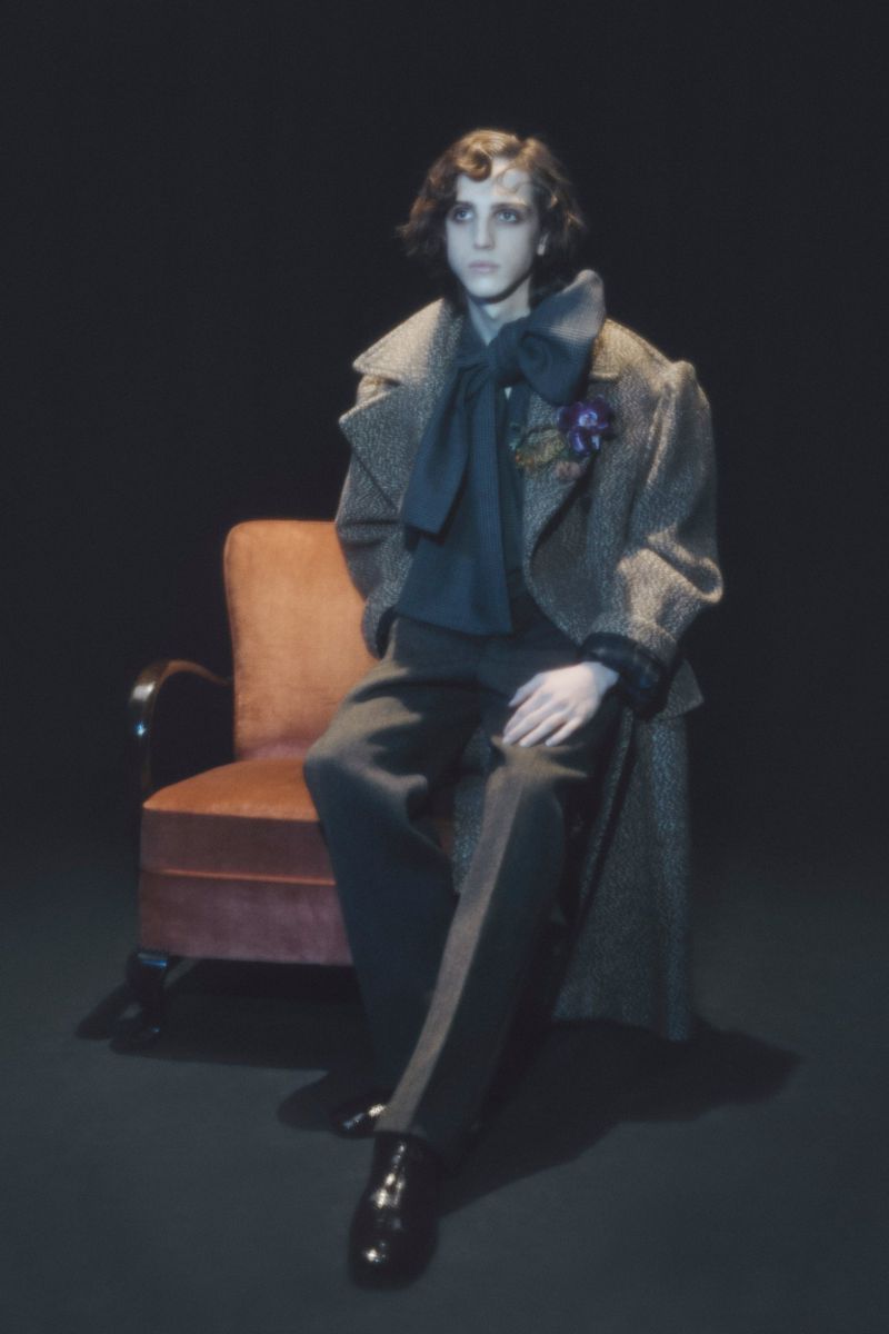 Elegant man in a patterned overcoat and dark trousers sits on a peach-colored chair against a solid black backdrop in a studio shot.
