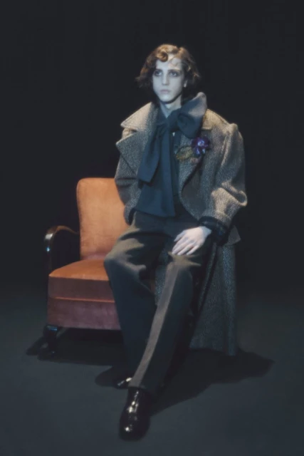 Elegant man in a patterned overcoat and dark trousers sits on a peach-colored chair against a solid black backdrop in a studio shot.