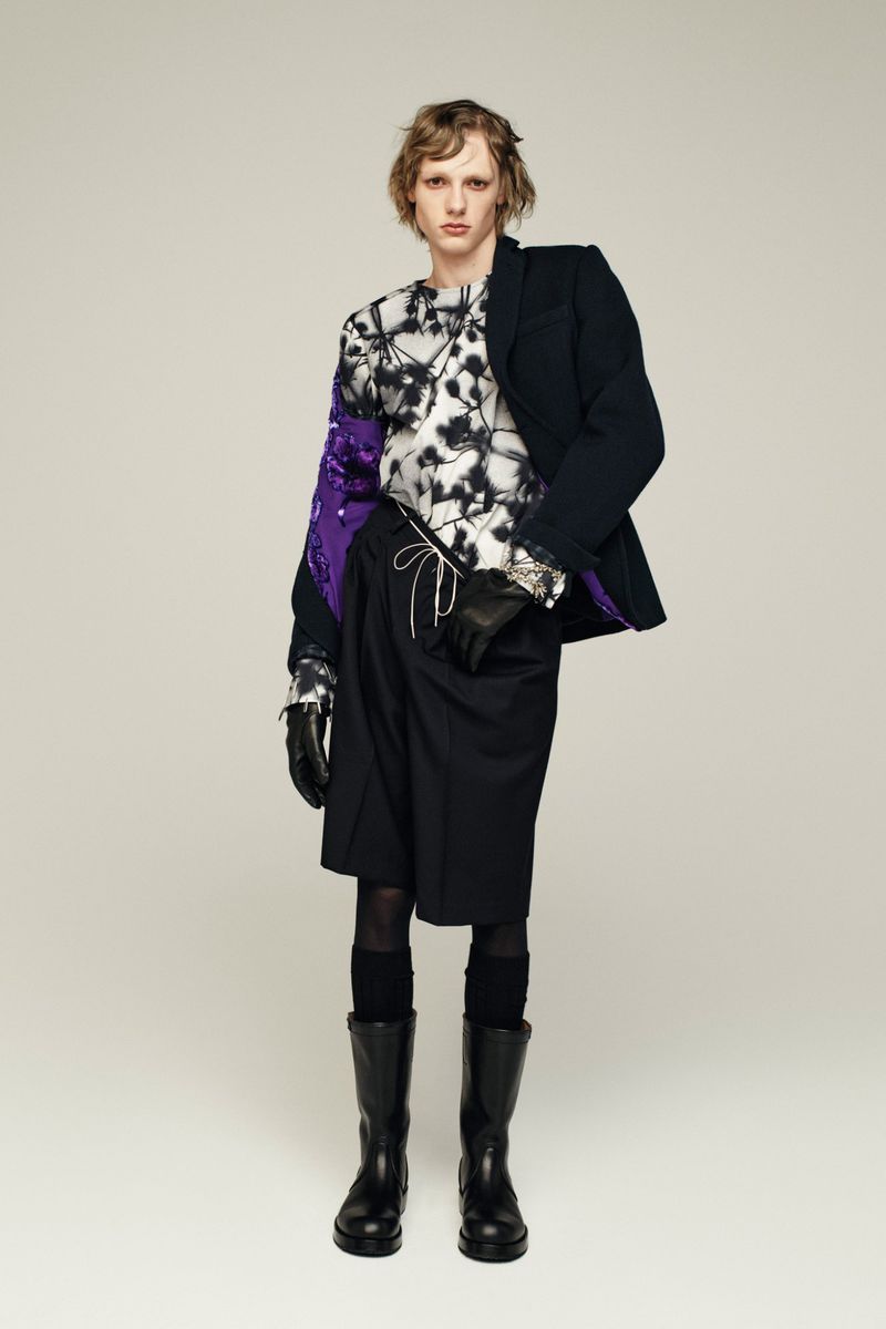 Male fashion model wears a graphic print top, black blazer, shorts, tights, boots, and gloves against a light background in a studio.