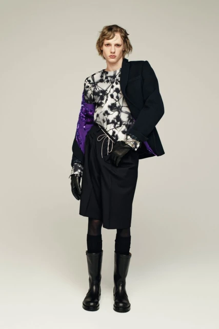 Male fashion model wears a graphic print top, black blazer, shorts, tights, boots, and gloves against a light background in a studio.