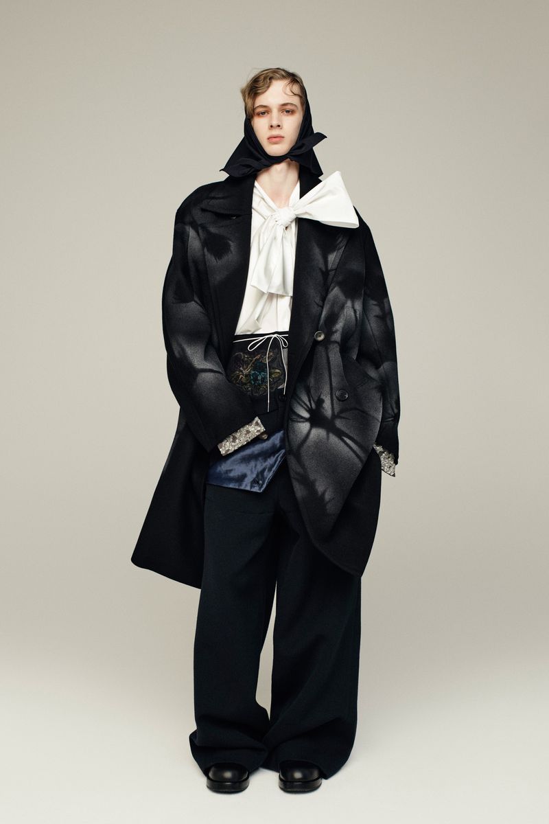 Fashion model in an oversized black coat, white ruffled shirt, and black pants poses in a studio with a neutral backdrop.