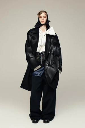 Fashion model in an oversized black coat, white ruffled shirt, and black pants poses in a studio with a neutral backdrop.