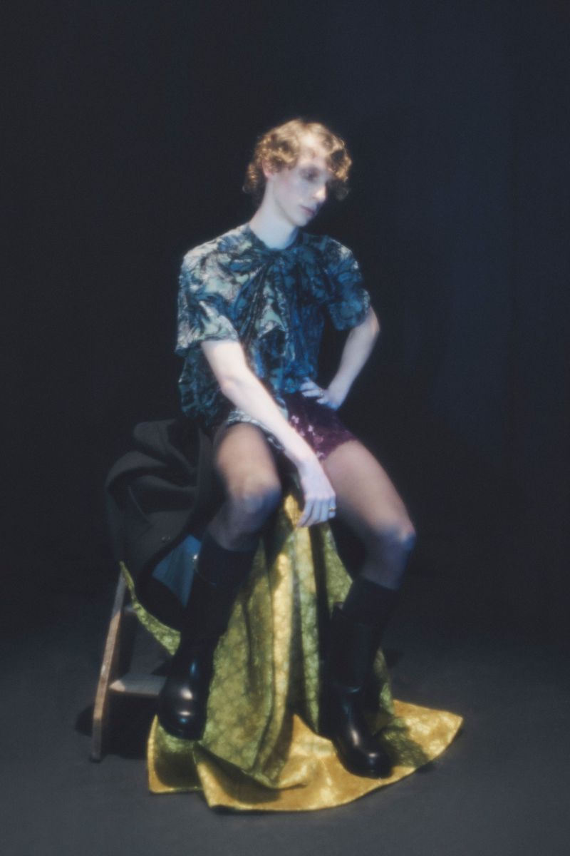 Young adult poses seated with black boots, patterned top and sequined short. Indoors against dark background, dramatic fashion portrait.