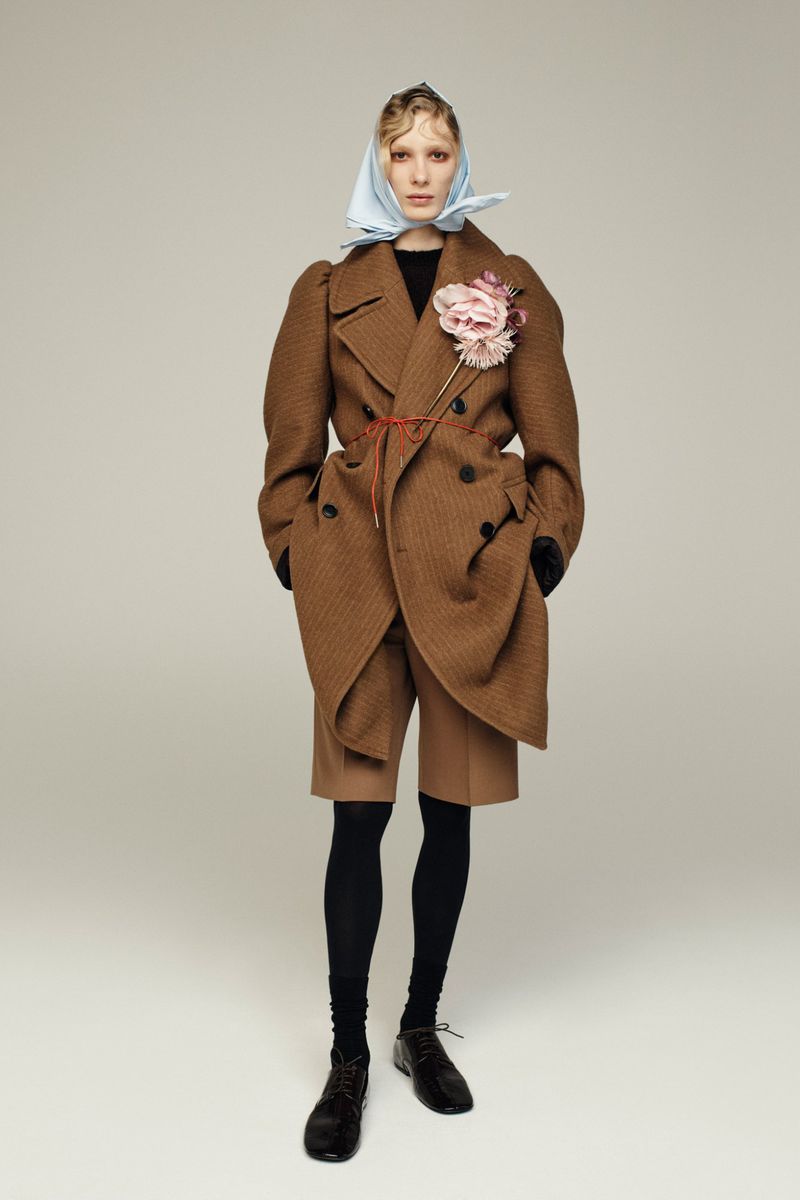 Full-shot fashion portrait of a woman wearing a brown overcoat, shorts, tights, and a headscarf in a bright studio setting.
